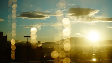 A cityscape is seen with a bright sun setting on the horizon, casting a warm glow over the scene. Light reflections create a bokeh effect with circular patterns, adding an artistic touch to the image. The sky is dotted with clouds, and silhouettes of buildings and streetlights are visible in the foreground.