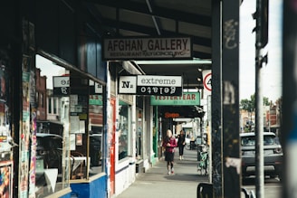 A street scene featuring various shops and commercial establishments. Signs for different businesses such as a restaurant named Afghan Gallery, a vintage clothing store, and an extreme tattoo parlor are visible. Two people are walking on the sidewalk, one closer to the foreground and the other in the distance. Several posters are visible on the storefront and the columns.