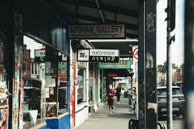 A street scene featuring various shops and commercial establishments. Signs for different businesses such as a restaurant named Afghan Gallery, a vintage clothing store, and an extreme tattoo parlor are visible. Two people are walking on the sidewalk, one closer to the foreground and the other in the distance. Several posters are visible on the storefront and the columns.
