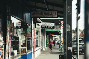 A street scene featuring various shops and commercial establishments. Signs for different businesses such as a restaurant named Afghan Gallery, a vintage clothing store, and an extreme tattoo parlor are visible. Two people are walking on the sidewalk, one closer to the foreground and the other in the distance. Several posters are visible on the storefront and the columns.