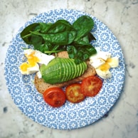 A plate with a slice of whole-grain bread topped with avocado slices, boiled eggs, cherry tomatoes, and spinach leaves, all arranged neatly. The plate has a blue and white geometric pattern, set against a marble background.