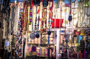 A vibrant collection of colorful necklaces, beads, and pendants is displayed, hanging from different levels. Various materials and styles are present, including metallic chains, beaded strands, and ornamental trinkets. The arrangement appears artistic and eclectic, with a mix of textures and colors creating an eye-catching display.