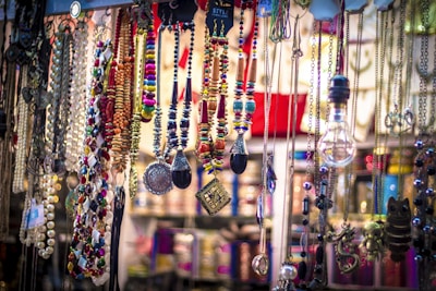A vibrant collection of colorful necklaces, beads, and pendants is displayed, hanging from different levels. Various materials and styles are present, including metallic chains, beaded strands, and ornamental trinkets. The arrangement appears artistic and eclectic, with a mix of textures and colors creating an eye-catching display.