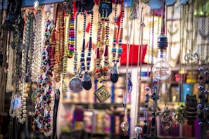 A vibrant collection of colorful necklaces, beads, and pendants is displayed, hanging from different levels. Various materials and styles are present, including metallic chains, beaded strands, and ornamental trinkets. The arrangement appears artistic and eclectic, with a mix of textures and colors creating an eye-catching display.