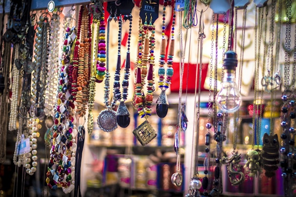 A vibrant collection of colorful necklaces, beads, and pendants is displayed, hanging from different levels. Various materials and styles are present, including metallic chains, beaded strands, and ornamental trinkets. The arrangement appears artistic and eclectic, with a mix of textures and colors creating an eye-catching display.