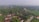 An aerial view of a village featuring a cluster of houses with brown roofs surrounded by dense greenery. There are various trees and plants scattered throughout the village. A winding road passes through the area, with a few vehicles visible. In the background, there are more trees that extend into the horizon.