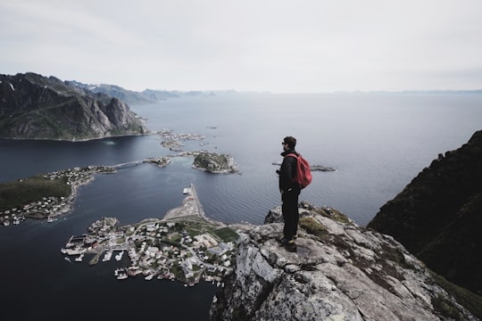 man standing on top of a cliff facing cityscape by water in Reine Norway