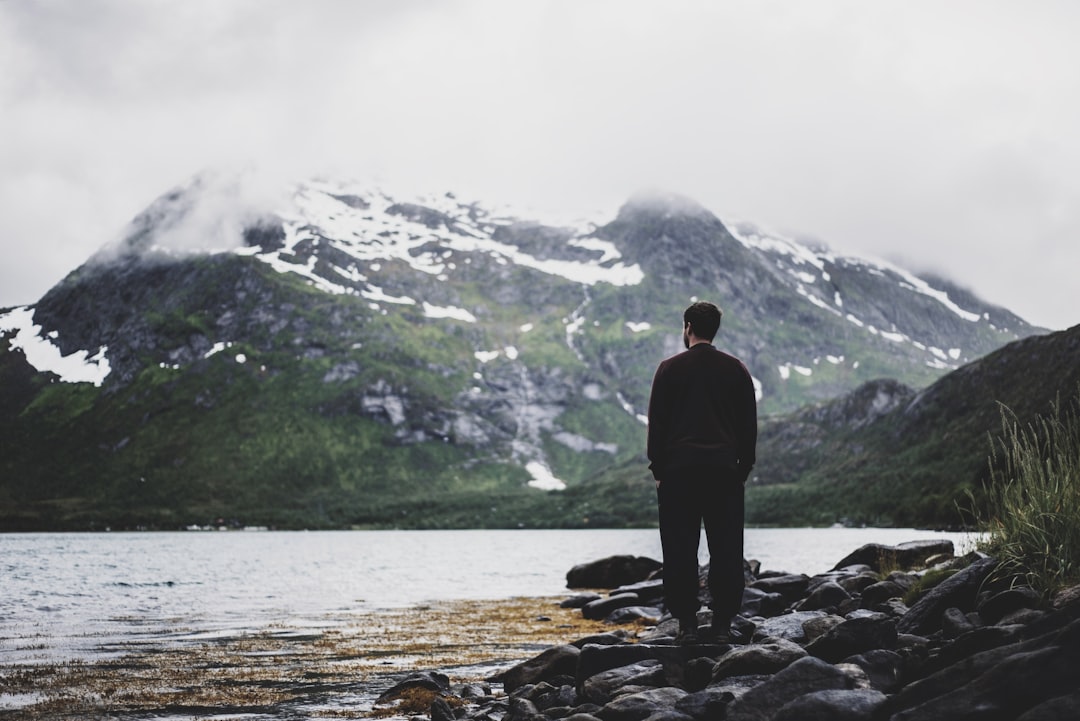 photo of man standing near seashore facing mountain covered with snow