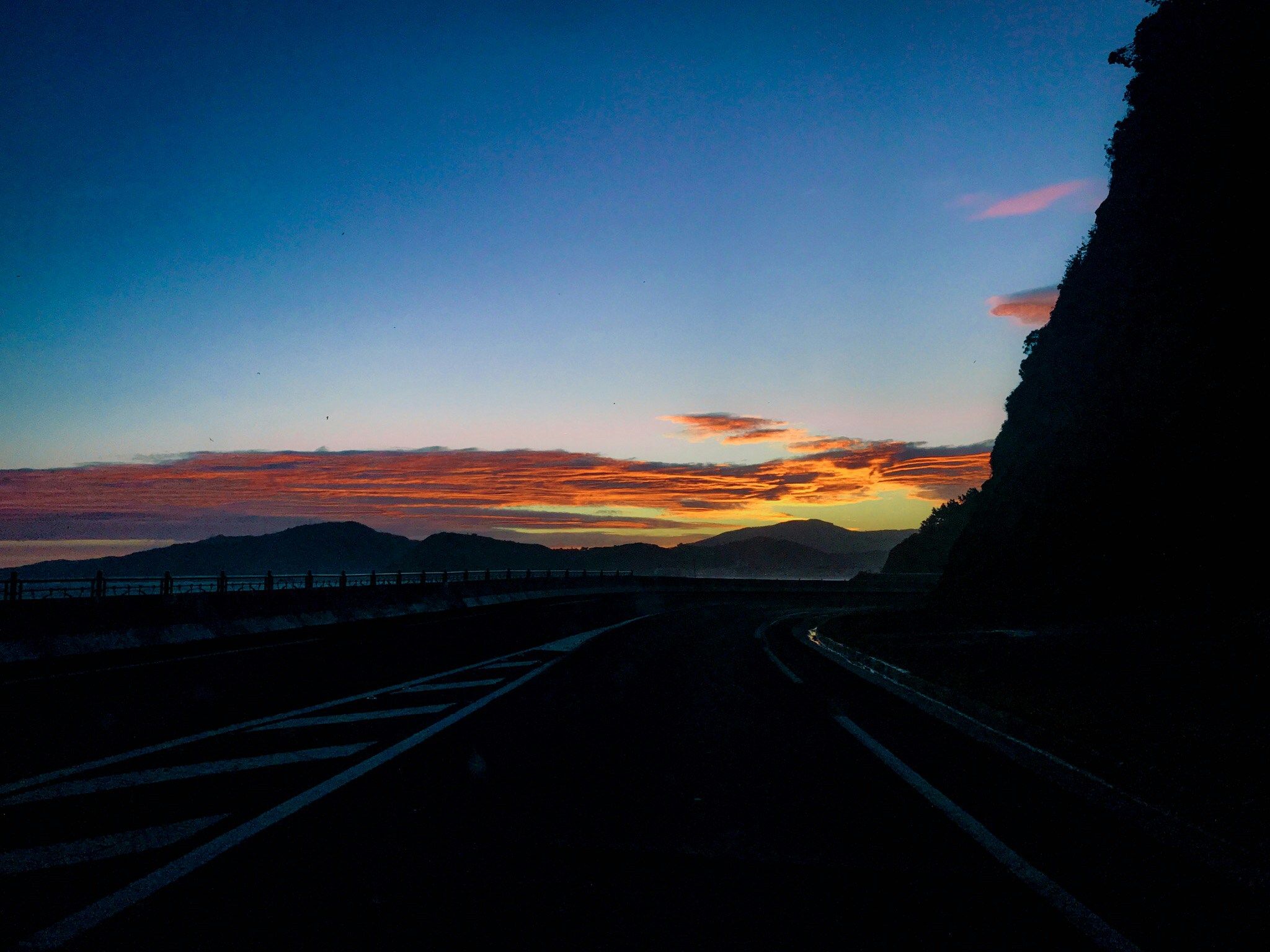 Winding mountain road at sunrise viewed from above, dark forest on both sides, orange-tinted light breaking through clouds, cinematic drone perspective