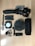A collection of videography and recording equipment is arranged on a wooden surface. The items include a handheld gimbal stabilizer, two rechargeable batteries, a compact LED light panel, a microphone with a windscreen cover, a small mount, and an Anker speaker.