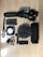 A collection of videography and recording equipment is arranged on a wooden surface. The items include a handheld gimbal stabilizer, two rechargeable batteries, a compact LED light panel, a microphone with a windscreen cover, a small mount, and an Anker speaker.