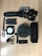A collection of videography and recording equipment is arranged on a wooden surface. The items include a handheld gimbal stabilizer, two rechargeable batteries, a compact LED light panel, a microphone with a windscreen cover, a small mount, and an Anker speaker.