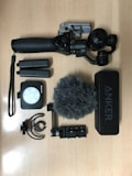 A collection of videography and recording equipment is arranged on a wooden surface. The items include a handheld gimbal stabilizer, two rechargeable batteries, a compact LED light panel, a microphone with a windscreen cover, a small mount, and an Anker speaker.