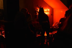 A dimly lit room featuring a neon sign with partially visible text. Silhouettes of people are gathered around low tables, creating a cozy, intimate atmosphere. The lighting casts a warm, orange-red glow which enhances the shadowy ambiance.