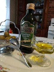 A bottle of Filippo Berio Extra Virgin Olive Oil is placed on a floral-patterned plate beside a small bowl of olives. A spoon rests against the bowl which is held by a silver holder. In the foreground, there is a glass dish containing olive oil. The background includes kitchen cabinets with plates displayed and a portion of a stove, creating a rustic kitchen setting.