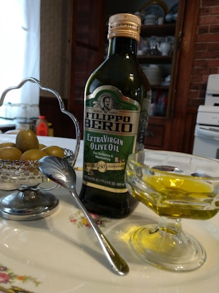 A bottle of Filippo Berio Extra Virgin Olive Oil is placed on a floral-patterned plate beside a small bowl of olives. A spoon rests against the bowl which is held by a silver holder. In the foreground, there is a glass dish containing olive oil. The background includes kitchen cabinets with plates displayed and a portion of a stove, creating a rustic kitchen setting.
