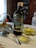 A bottle of Filippo Berio Extra Virgin Olive Oil is placed on a floral-patterned plate beside a small bowl of olives. A spoon rests against the bowl which is held by a silver holder. In the foreground, there is a glass dish containing olive oil. The background includes kitchen cabinets with plates displayed and a portion of a stove, creating a rustic kitchen setting.