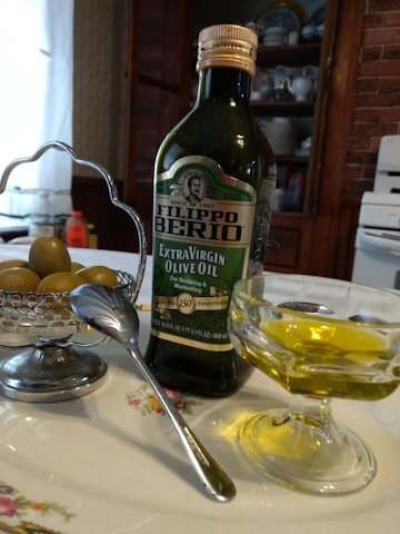 A bottle of Filippo Berio Extra Virgin Olive Oil is placed on a floral-patterned plate beside a small bowl of olives. A spoon rests against the bowl which is held by a silver holder. In the foreground, there is a glass dish containing olive oil. The background includes kitchen cabinets with plates displayed and a portion of a stove, creating a rustic kitchen setting.