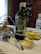 A bottle of Filippo Berio Extra Virgin Olive Oil is placed on a floral-patterned plate beside a small bowl of olives. A spoon rests against the bowl which is held by a silver holder. In the foreground, there is a glass dish containing olive oil. The background includes kitchen cabinets with plates displayed and a portion of a stove, creating a rustic kitchen setting.