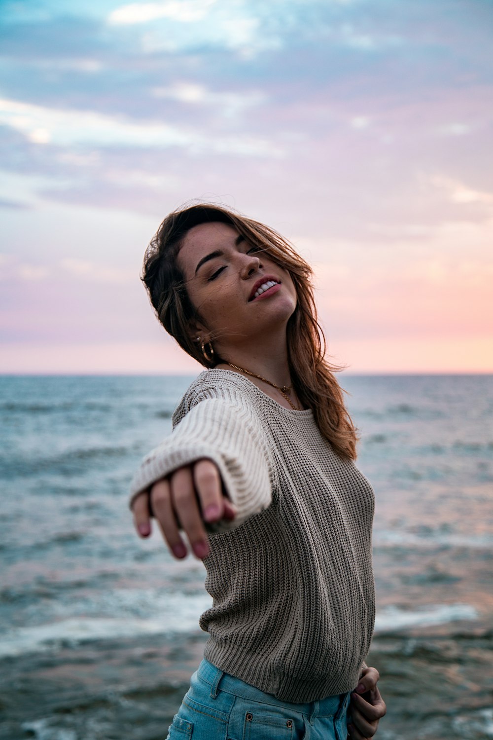 woman in grey sweater and blue denim bottoms reaching out arms and slightly looking up at beach under pink and blue sky
