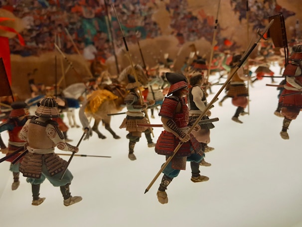An intricate scene of miniature samurai figures engaged in battle, each wearing traditional armor and wielding various weapons such as spears and swords. The figures are intricately detailed, and the scene recreates a dynamic and chaotic battle scenario with multiple figures in motion.