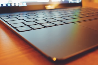 A close-up angle of a laptop keyboard, highlighting the keys and the trackpad. The lighting is warm, casting a soft glow over the black surface of the keyboard. The focus is on the edge, emphasizing the sleek and modern design.