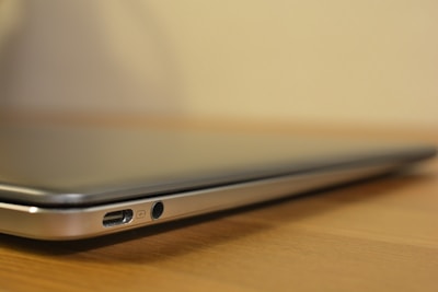A close-up view of a slim laptop or notebook computer, showing the side ports including USB and audio jack. The laptop is resting on a wooden surface with a blurred background.