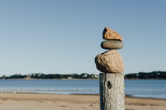 A stack of three stones is carefully balanced on a wooden post, set against a serene background of a sandy beach and calm body of water. The horizon is dotted with darker foliage, and the sky is clear and blue.