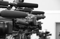 A close-up view of a row of professional television cameras equipped with large microphones, all lined up and focused in the same direction, suggesting a press conference or media event.