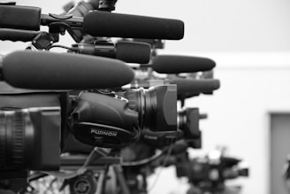 A close-up view of a row of professional television cameras equipped with large microphones, all lined up and focused in the same direction, suggesting a press conference or media event.