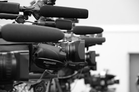 A close-up view of a row of professional television cameras equipped with large microphones, all lined up and focused in the same direction, suggesting a press conference or media event.