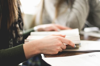 A close-up of a person holding a book while engaging in reading or study. Another person is sitting nearby, both seemingly focused on their tasks. The setting suggests a studious or collaborative environment, possibly at a library or study room.
