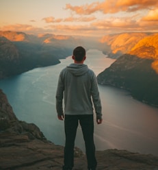 man wearing grey hoodie standing on mountain looking at river