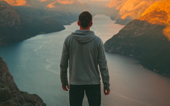 man wearing grey hoodie standing on mountain looking at river