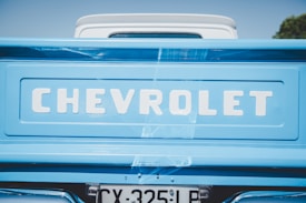 The image depicts the rear end of a Chevrolet vehicle, showcasing the brand name prominently on a light blue background. The word 'Chevrolet' appears in bold, white letters. Below the brand name is a portion of a license plate with the registration number partially visible. The background is mostly clear with a hint of greenery on one side.