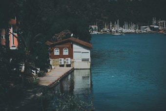 A tranquil lakeside scene features a small house built partially over the water with a red roof and white walls. Air conditioning units are visible on the side of the house. The water reflects the surrounding greenery and buildings. In the background, numerous sailboats are docked at a marina. The area is lush with dense forest and a paved walkway leads to the house.