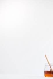 A minimalistic composition featuring a clear glass jar filled with honey placed on a white surface. A wooden honey dipper rests inside the jar. The background is bright and uncluttered, accentuating the simplicity of the scene.