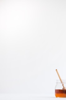 A minimalistic composition featuring a clear glass jar filled with honey placed on a white surface. A wooden honey dipper rests inside the jar. The background is bright and uncluttered, accentuating the simplicity of the scene.