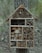 A wooden insect house is attached to a tree trunk. The house is divided into various compartments filled with natural materials like bamboo tubes, wooden blocks, and twigs, offering a habitat for insects. The background consists of a blurred forest with trees and greenery.