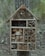 A wooden insect house is attached to a tree trunk. The house is divided into various compartments filled with natural materials like bamboo tubes, wooden blocks, and twigs, offering a habitat for insects. The background consists of a blurred forest with trees and greenery.