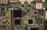 A detailed view of a computer motherboard with various integrated circuits and components including chips, capacitors, and connectors. The board is densely populated with intricate pathways and electronic parts, indicative of complex technology.