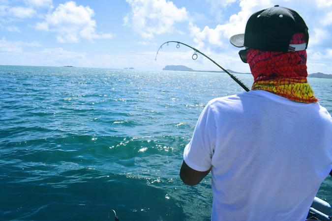 Angler seen from behind on a boat, wearing a black cap and a red and yellow patterned gaiter pulled up over the face, rod bent over open blue water