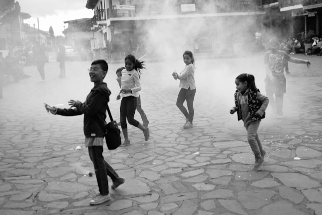 Children are joyfully playing and running on a cobblestone street, surrounded by a cloud of smoke or dust, with buildings and people in the background.