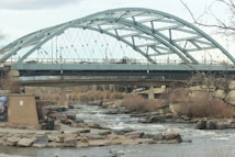 A large, arched steel bridge spans across a flowing river with rocks lining the riverbanks. Bare trees and shrubs are visible, indicating a likely winter or early spring setting. The bridge has a distinctive geometric design with cables and supports visible. An expansive view of the sky and some distant structures or trees is seen in the background.