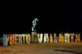 A colorful display of large, decorated letters spelling out 'Puerto Vallarta' with a seahorse statue perched on top of a stone pedestal. The letters feature various artistic designs and illustrations, representing cultural elements. The scene is set against a dark background, highlighting the vibrant colors of the artwork.