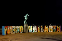 A colorful display of large, decorated letters spelling out 'Puerto Vallarta' with a seahorse statue perched on top of a stone pedestal. The letters feature various artistic designs and illustrations, representing cultural elements. The scene is set against a dark background, highlighting the vibrant colors of the artwork.