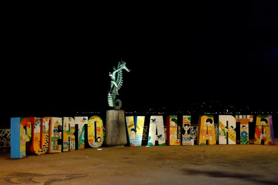 A colorful display of large, decorated letters spelling out 'Puerto Vallarta' with a seahorse statue perched on top of a stone pedestal. The letters feature various artistic designs and illustrations, representing cultural elements. The scene is set against a dark background, highlighting the vibrant colors of the artwork.