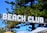 A sign with the words 'BEACH CLUB' in bold, white letters is prominently displayed against a backdrop of tall green trees and a vivid blue sky. Below the sign, striped umbrellas in shades of blue and white are visible along with string lights that add a festive ambiance.