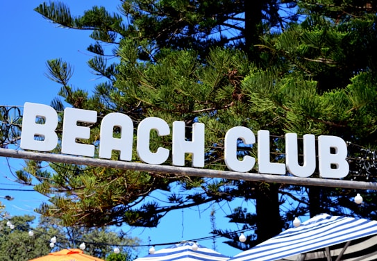 A sign with the words 'BEACH CLUB' in bold, white letters is prominently displayed against a backdrop of tall green trees and a vivid blue sky. Below the sign, striped umbrellas in shades of blue and white are visible along with string lights that add a festive ambiance.