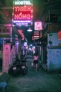 A narrow, dimly lit urban alley with colorful neon signs advertising hostels and guest houses. A person is walking away carrying a small bag, and there are parked motorcycles along the alley.